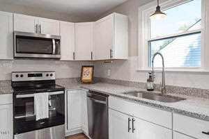Modern kitchen with white cabinets, stainless steel appliances including a stove, microwave, and dishwasher, granite countertops, a double sink under a window, and a framed picture on the counter.