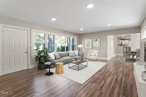 A spacious living room with wood flooring, beige walls, a gray sectional sofa, a glass coffee table, a light armchair, large windows, and wall art. The room opens to a kitchen and dining area.