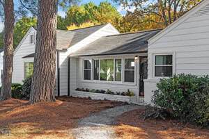Single-story white house with black trim, large front windows, a black front door, and a small garden bed. Pine needles cover the ground, and two large trees stand near the entrance. Shrubs line the house's exterior.