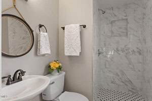 A bathroom with white marble tile shower, round mirror above a white sink, white toilet with a flower arrangement, and two white patterned towels on black towel bars.