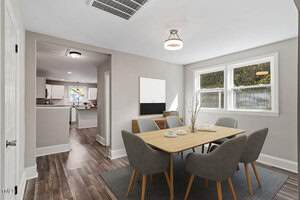 A dining room with a wooden table and four gray chairs on a rug; minimal decor on the table. Large windows let in natural light. In the background, there is a view into a white kitchen with wood-style flooring.