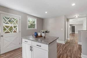 A kitchen area with a small island featuring a granite countertop and white cabinets, adjacent to a door with a diamond-pane window, wood flooring, and neutral-colored walls.
