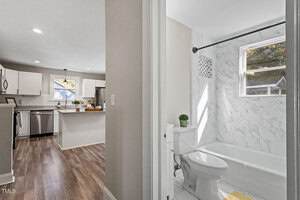 View from a hallway showing a kitchen with white cabinets, stainless steel appliances, and wood flooring on the left, and a bathroom with a bathtub, toilet, marble tile walls, and a window on the right.