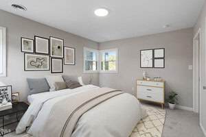 A bedroom with a neatly made bed, neutral bedding, a gallery wall above the headboard, a white dresser with decor, a small plant, and two windows letting in natural light.