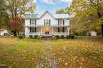 A two-story white house with a green metal roof, wraparound porch, and steps leading to the front door sits at 1734 Rocky Ford Road in Kittrell, surrounded by autumn trees and a grassy yard dotted with fallen leaves.