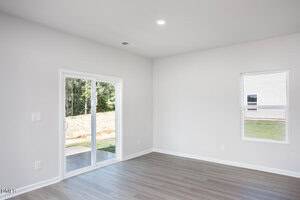 A bright, empty room with light gray walls and wood-style flooring features a sliding glass door leading outside and a single window. The space is well-lit with natural light.