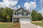 Two-story Oxford-style suburban house with gray siding, white trim, and a double garage at 1114 Rhino Bend. The home sits on a newly landscaped lawn with a young tree, bordered by woods and a partly cloudy sky above.
