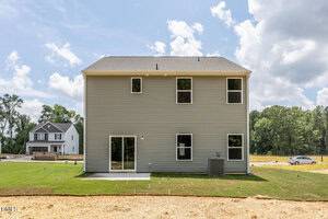 A two-story house with light gray siding, five rectangular windows, a sliding glass door, an exterior air conditioning unit, and a small concrete patio, with another house and trees visible in the background.