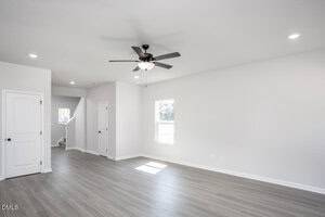 A bright, empty room with light gray wood flooring, white walls, a ceiling fan with a light, two white doors, one window, and a small stairway visible in the background.