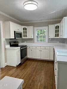 A kitchen with white cabinets, stainless steel appliances, a double sink, and a window above the counter. The floor is wood, and there is a round ceiling light. The walls are light and the backsplash is gray tile.