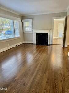 A bright, empty living room with hardwood floors, cream-colored walls, a black fireplace with a white mantel, two large windows with blinds, and an open doorway leading to another room.