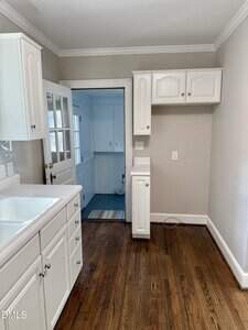 A kitchen with white cabinets, a white sink, and brown hardwood floors. An open door leads to a smaller adjoining room with white cabinetry and a window. The walls are painted light beige.
