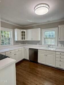 A modern kitchen with white cabinets, white countertops, stainless steel dishwasher, double windows above the sink, and wood flooring. The space is well-lit by a round ceiling light.