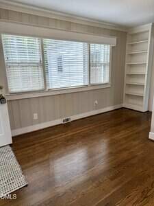 A room with hardwood floors, light-colored paneled walls, three large windows with blinds, a built-in white bookshelf, and a white door partially visible on the left.