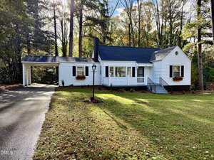 A single-story white house with black shutters and a covered carport on the left, 1609 Cypress Drive in Henderson features a front porch, two window boxes, and is surrounded by trees and a grassy yard with a paved driveway.