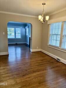 A room with hardwood floors, beige walls, and white trim, featuring a chandelier and large windows. An arched doorway leads to a second room with blue walls and more windows.