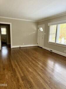 Empty living room with hardwood floors, beige walls, white trim, a front door with a window, a large front-facing window, and an open doorway leading to another room in the background.