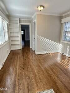 A bright, empty room with hardwood floors, built-in white shelves, beige walls, and ample natural light from multiple windows. The room has a ceiling light fixture and an open doorway leading to another area.