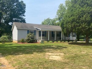 A single-story, light gray house with blue shutters and a front porch sits on a grassy lawn at 910 Vanco Mill Road in Henderson, surrounded by trees and landscaping with shrubs and mulch beds in front.