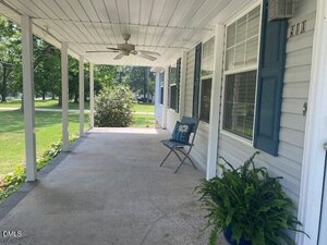 A covered front porch with a ceiling fan, a single blue chair with a pillow, a potted fern, blue shutters, and white siding. The porch overlooks a grassy yard with trees and bushes.