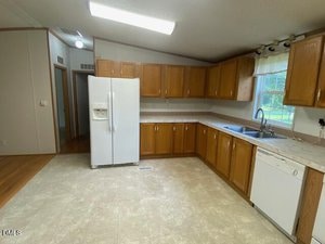 A kitchen with wooden cabinets, white countertops, a white refrigerator, dishwasher, double sink, and a window above the sink. The floor is light-colored tile, and the lighting is fluorescent.
