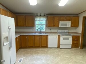 A kitchen with wood cabinets, white appliances including a refrigerator, dishwasher, stove, and microwave, a double sink under a window with a white curtain, and overhead fluorescent lighting.