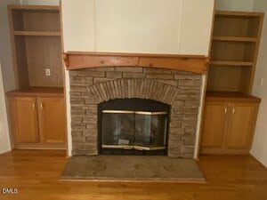 A stone fireplace with a wood mantel is centered between two built-in wooden shelves and cabinets. The floor is hardwood, and the walls are light-colored.