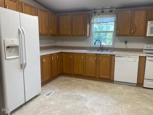 A kitchen with wood cabinets, white appliances including a refrigerator, dishwasher, and stove, a double sink under a window with a curtain, and light-colored tile flooring. The counters are uncluttered.