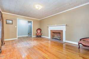 A living room with light brown walls, a wooden floor, a white fireplace, two vintage pink chairs, and a picture on the wall. The room is brightly lit with a ceiling light and has a doorway leading to another area.