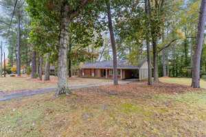 A single-story brick house with a covered front porch, surrounded by tall trees and a large, grassy yard with scattered leaves. A paved driveway leads to the front of the home.