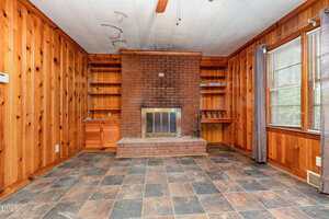 A wood-paneled room with a brick fireplace centered on one wall, flanked by built-in wood shelves and cabinets. The floor is tiled in a dark, mottled pattern. Large windows with curtains are on the right side.