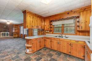 A spacious kitchen with wood-paneled cabinets, a double sink under a window, tiled floors, and open shelving. The adjoining room features a fireplace, wood walls, and large windows.