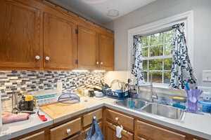 A kitchen with wooden cabinets, a tiled backsplash, and a double sink under a window with floral curtains. Countertops hold various dishes, a cutting board, and utensils. A water stain is visible on the ceiling.