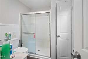 A bathroom with a glass-enclosed shower, a white toilet, cleaning supplies on the toilet tank, and a closed white door. The walls are light-colored and the floor is dark.