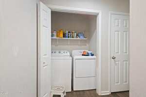 A laundry area with a washing machine and dryer beneath a white shelf holding detergent and cleaning supplies. The space is enclosed by open double doors, with a small white step stool in front.