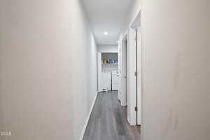 A hallway with light-colored walls and dark flooring leads to a small laundry area with a washer and dryer. There are shelves above the machines holding various items. Two doorways are on the right side of the hallway.