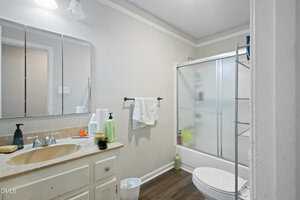 A bathroom with a beige countertop, white sink, and mirror above. Towels hang on a black towel rack. There is a white toilet, shelving unit, frosted glass shower door, and wood-style flooring. Toiletries are on the counter.