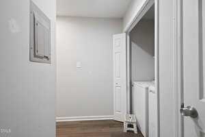 A small laundry area with a top-loading washing machine, a small white step stool, wood-look flooring, a wall-mounted electrical panel, and a partially open closet door. The walls are plain and light-colored.
