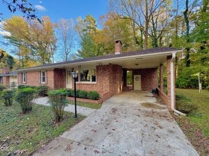 A single-story brick house with a carport on the right, a front yard with trimmed bushes and a lamp post, and trees with autumn leaves in the background. The driveway is concrete and partially covered.