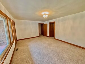 Unfurnished room with beige carpet, white walls, a ceiling light fixture, two wooden doors, a large window, and natural light coming through the window.