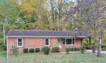 Charming single-story brick house at 105 Sunset Circle, Henderson, featuring a covered front porch, large front window, and dark shingle roof. Bushes line the home and a tree graces the lawn, with tall autumn trees in the background.
