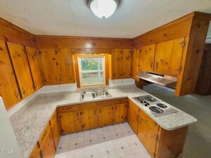 A small kitchen with wooden cabinets, a double sink beneath a window, a four-burner electric stovetop, granite countertops, tile flooring, and a ceiling light fixture. A range hood is above the stovetop.