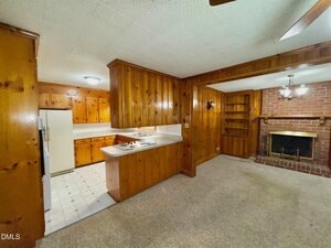 Wood-paneled room with carpeted floor, built-in shelves, and a brick fireplace. An open counter connects the space to a kitchen with white appliances and tile flooring. Ceiling lights illuminate both areas.