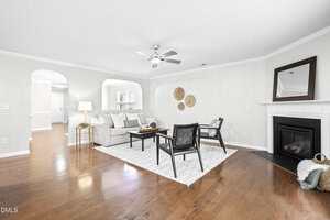 A spacious living room with hardwood floors, neutral walls, a white sofa, two black chairs, a fireplace with a mirror above it, and a white rug. Arched doorways lead to other rooms; a lamp and wall decor complete the space.
