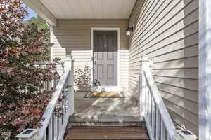 A front porch with beige siding, a gray door, white railing, a welcome mat, and a small outdoor light fixture next to the door. A leafy plant is on the left side of the entrance.