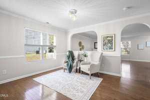 Bright sitting area with two beige armchairs, a small round table, a green throw blanket, and a patterned rug on wood floors. Large window, framed artwork, and arched doorways lead to adjacent rooms.