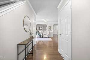 A hallway with wooden floors leads to a living room area. The hallway has a console table with a round mirror above it, and the living room features two chairs, a coffee table, and framed art on the walls.
