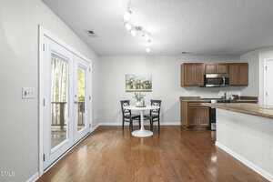 A kitchen and dining area with wooden floors, brown cabinets, and stainless steel appliances. A small round table with two chairs is set by the wall, and double glass doors lead to an outdoor space.