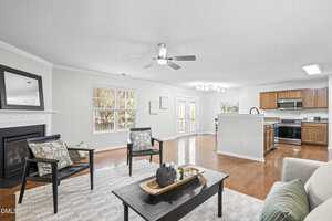 A spacious open-concept living room and kitchen featuring hardwood floors, a fireplace, neutral walls, wooden cabinets, stainless steel appliances, and large windows letting in natural light.