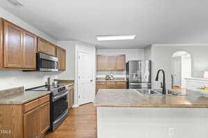 A modern kitchen with wood cabinets, stainless steel appliances, a double sink with a black faucet, beige countertops, and a walk-in pantry. The kitchen opens to a bright living area through an arched doorway.
